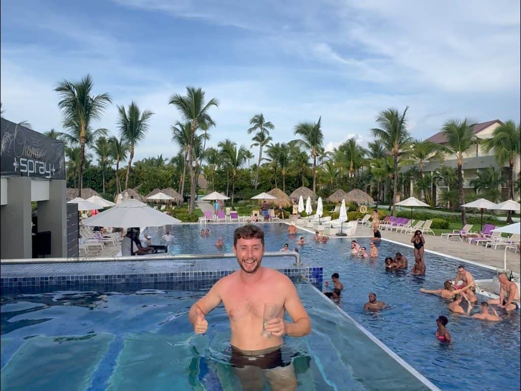 Relaxed man in a pool at a tropical resort with palm trees and other guests swimming.