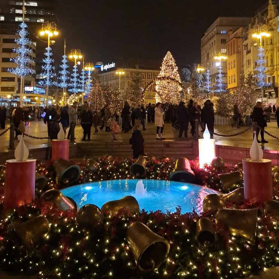 Festive Christmas market square with decorated trees, lights, and crowd at night.