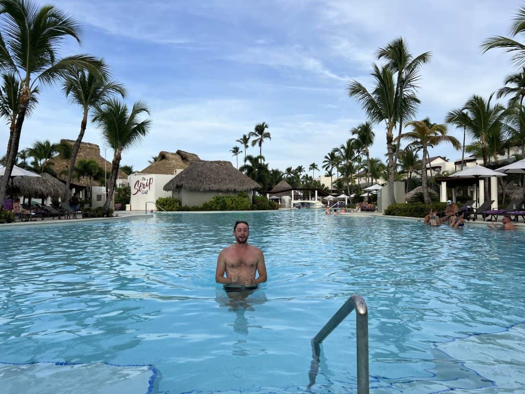 Relaxing man enjoying a tropical pool surrounded by palm trees and thatched roofs.