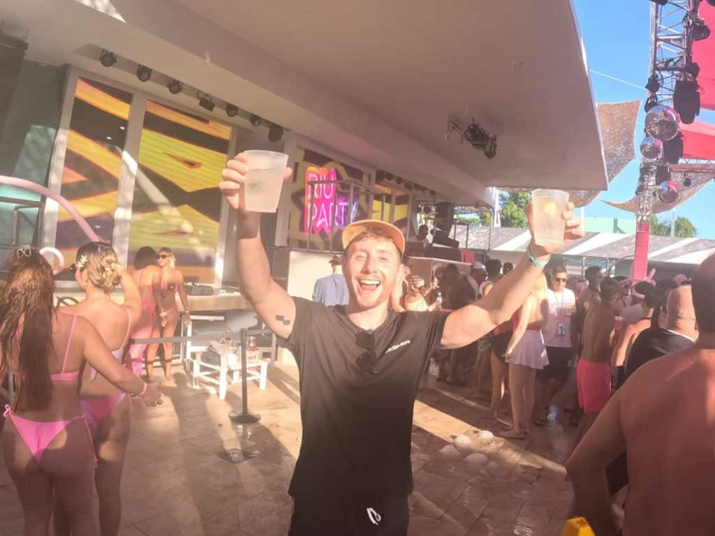 Vibrant pool party scene with cheerful man holding drinks in the air, lively crowd, and colorful lighting.