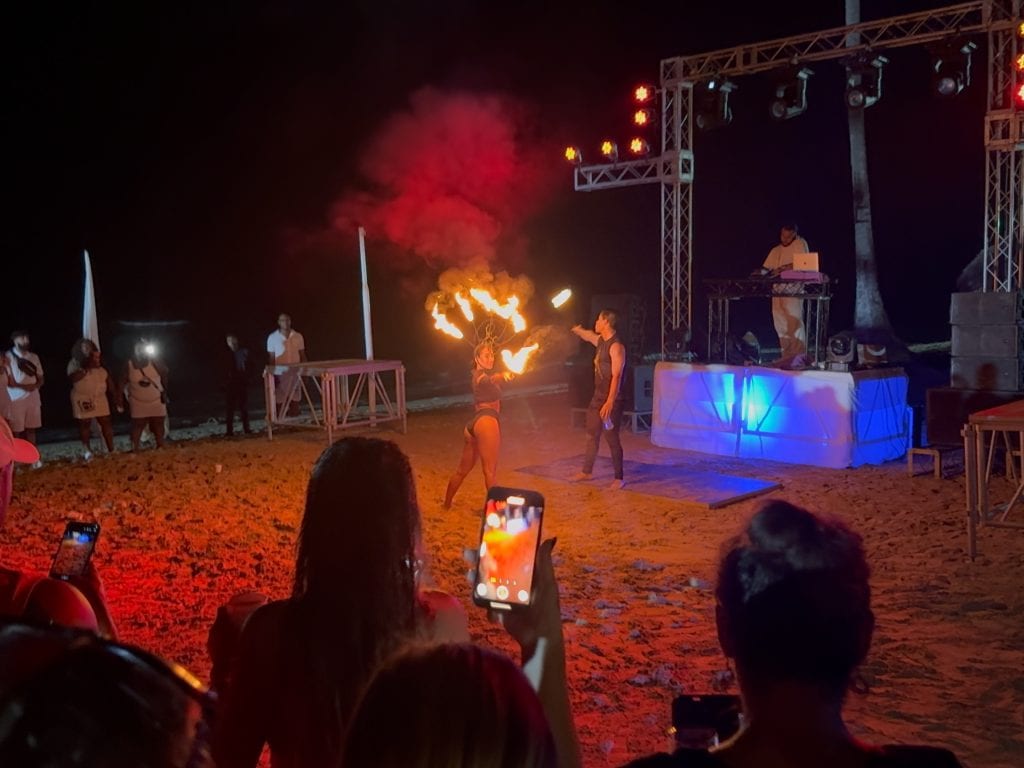 Vibrant fire dance performance on the beach at night with a DJ and audience watching.