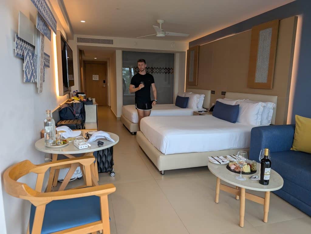Modern hotel room with two beds, sitting area, and a man holding a drink in a tropical resort.