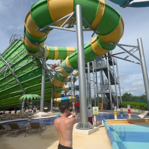 A man in swimwear at the colorful water park with large green and yellow slides.