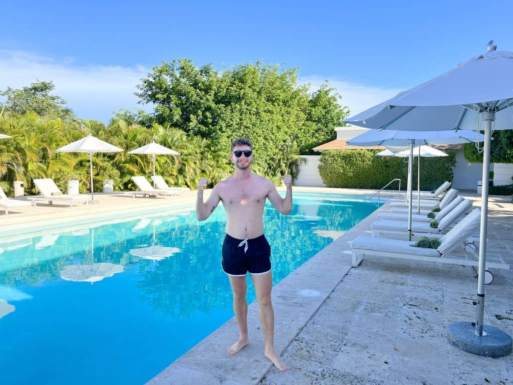 Relaxed man in swim shorts and sunglasses by the poolside with umbrellas and lush greenery.