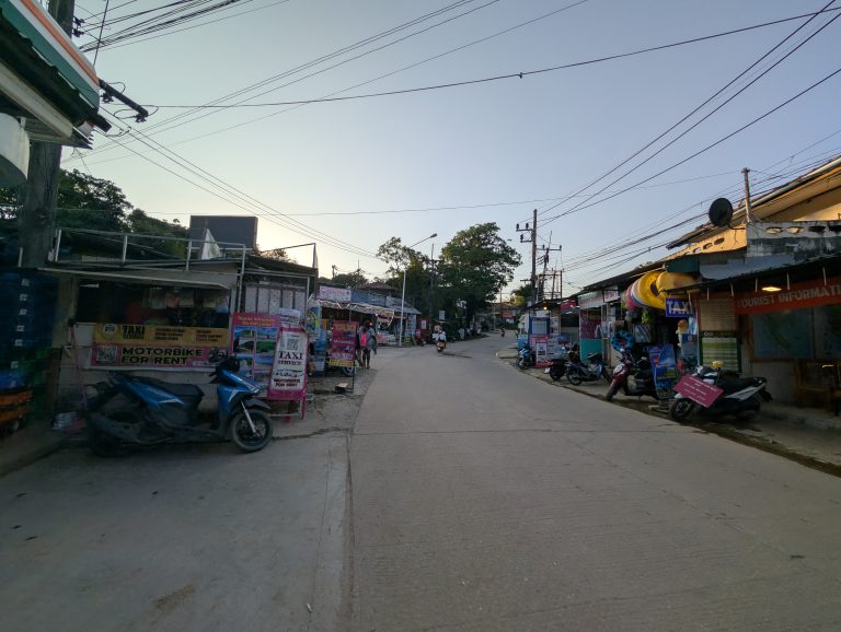 La carretera principal de Koh Lanta al atardecer, con motos aparcadas y un cartel de alquiler de motos