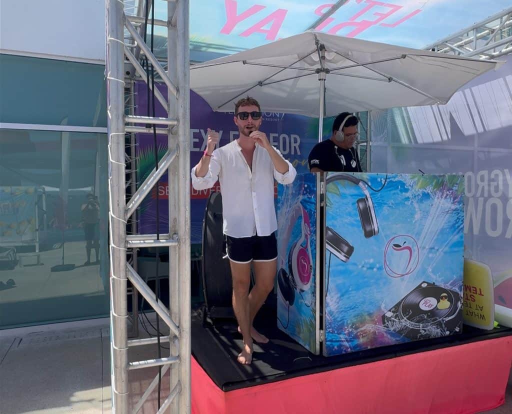 Male DJ performing at an outdoor beach event with a colorful promotional booth.