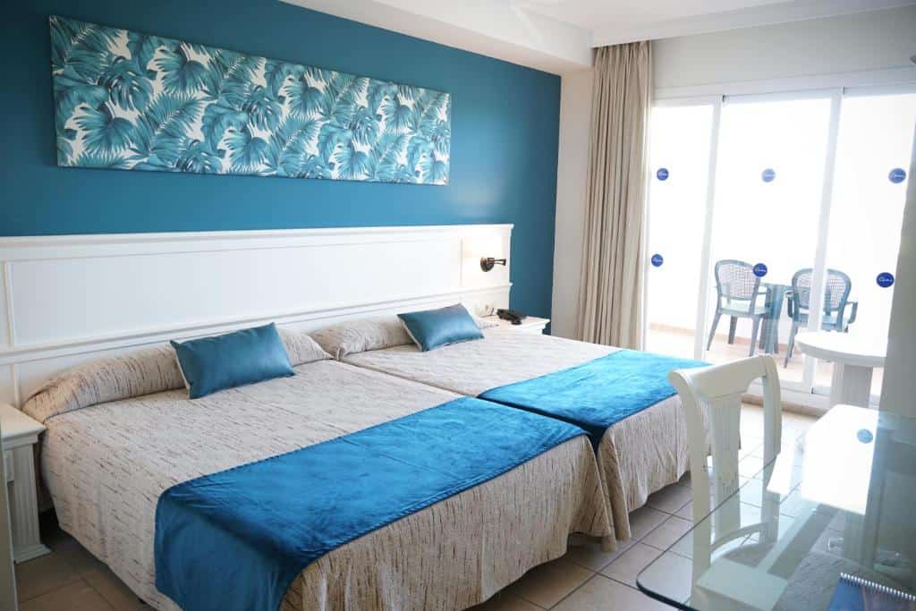 Bright hotel room with twin beds, blue accents, and balcony overlooking the sea.