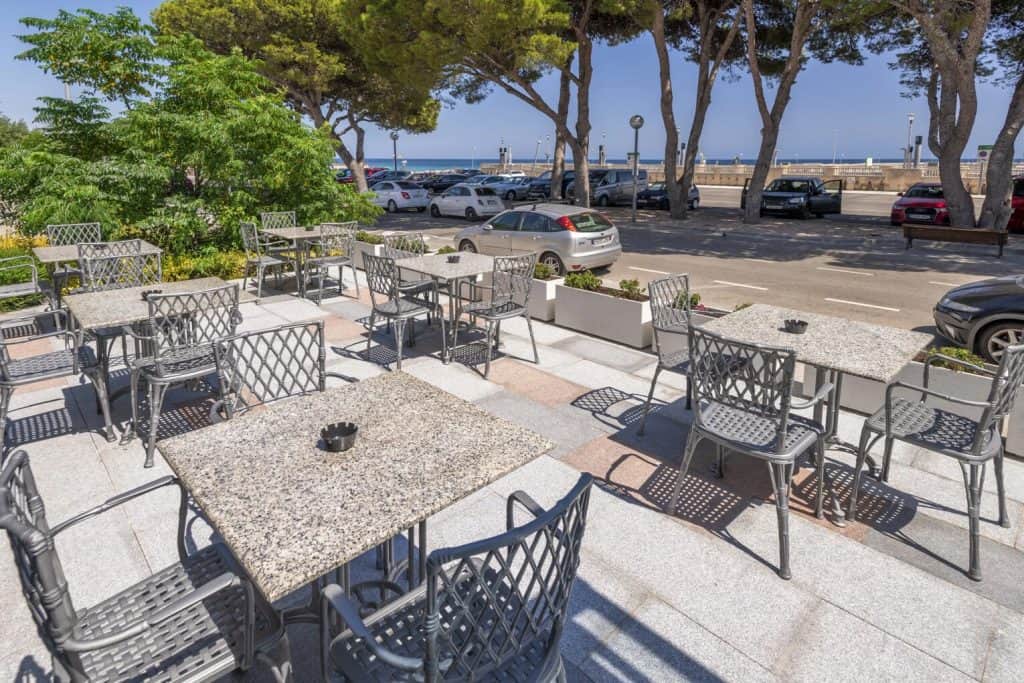 Sea view outdoor cafe with tables and chairs near parking lot and the ocean in the background.