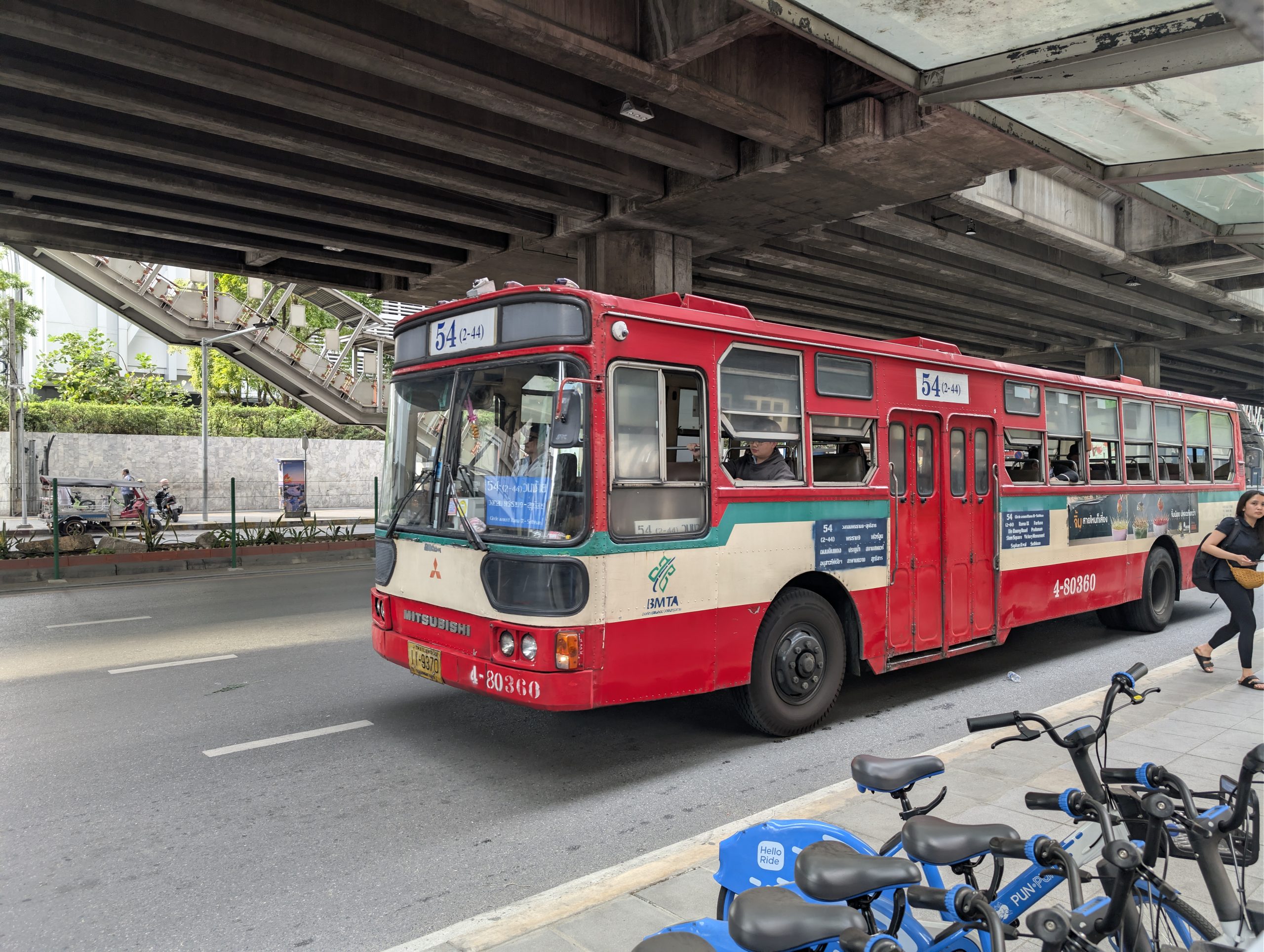 Autobús urbano rojo de la línea 54 circulando por Bangkok bajo la estructura elevada del BTS, con bicicletas de alquiler en primer plano