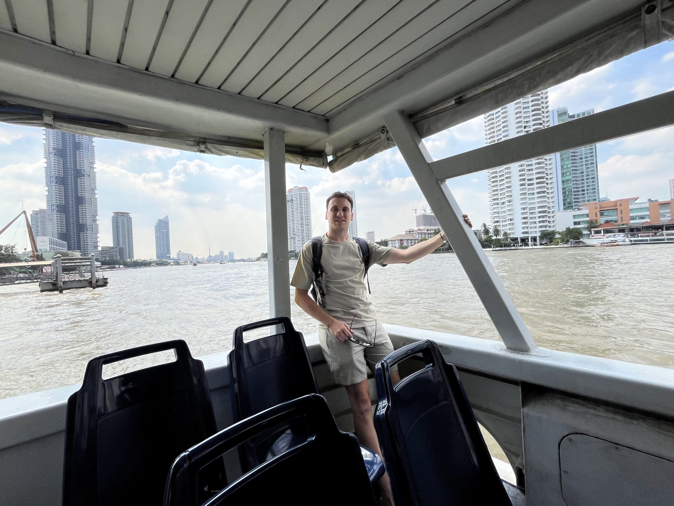 Víctor de pie en la cubierta de un barco público del río Chao Phraya en Bangkok, con los rascacielos de la orilla al fondo