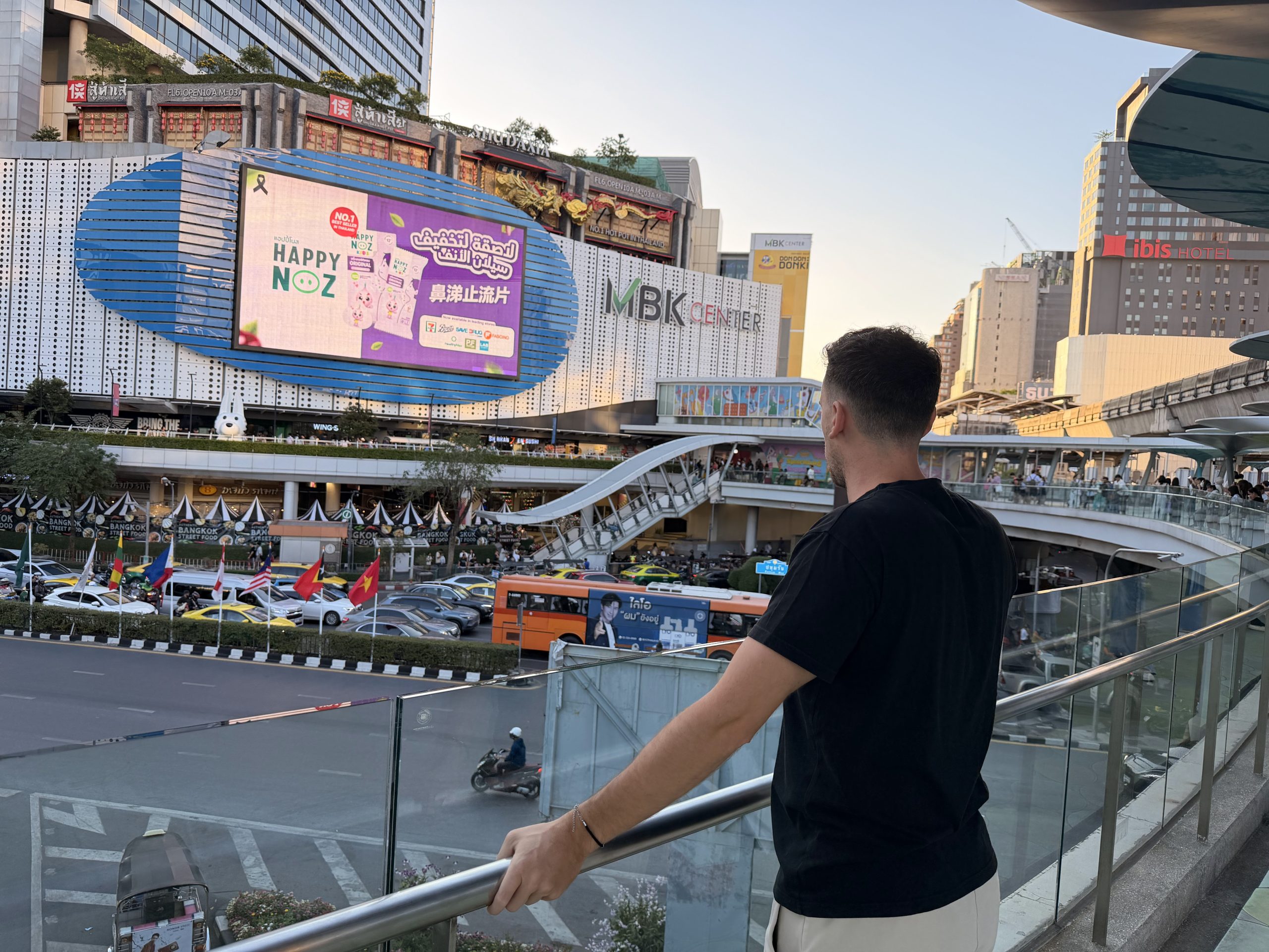 Víctor apoyado en la barandilla de una pasarela elevada frente al MBK Center de Bangkok, con el tráfico y las pantallas publicitarias