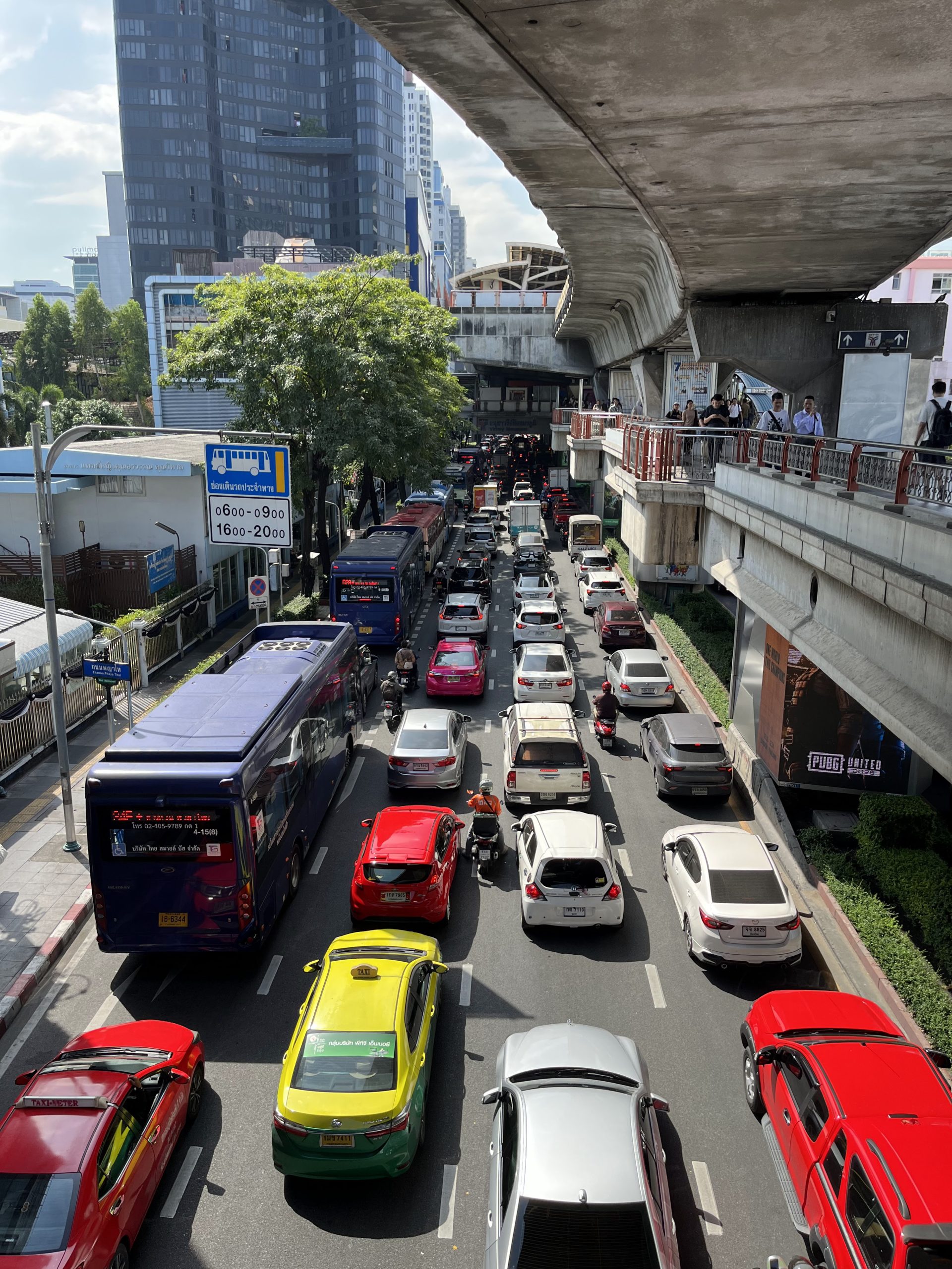 Atasco de coches, autobuses y motos en una avenida de Bangkok visto desde una pasarela elevada, con la estructura del BTS al lado