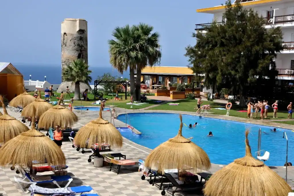 Relaxing hotel pool area with straw umbrellas, lounge chairs, and a historic tower in the background.