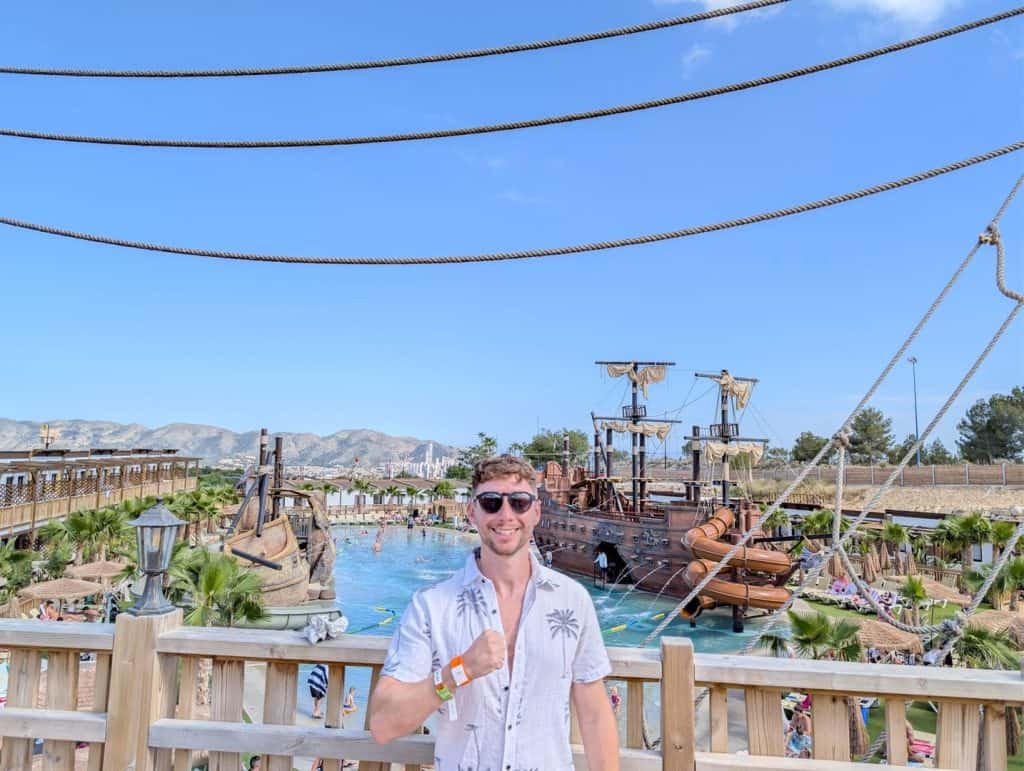 A young man with sunglasses at a pirate-themed water park with ships and water slides in the backgro.