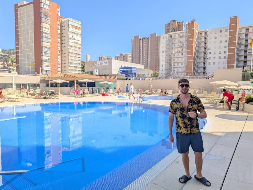 A man with sunglasses and a floral shirt standing by a swimming pool in an urban hotel setting.