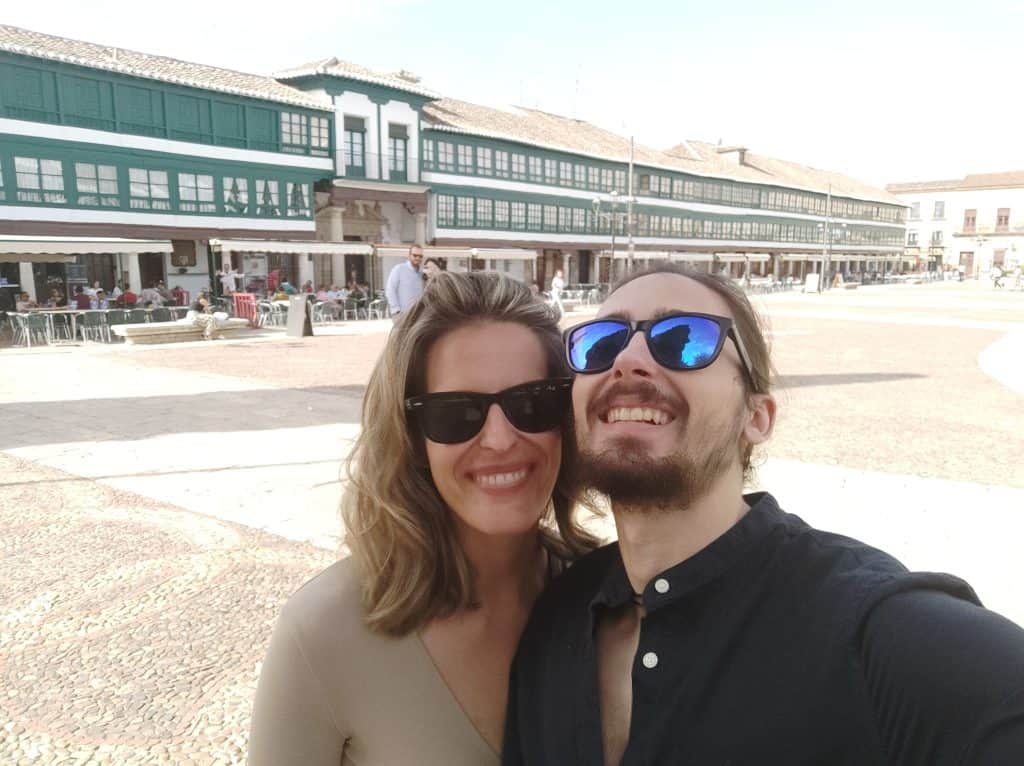 A smiling couple taking a selfie in a lively outdoor plaza with historic buildings in the background.