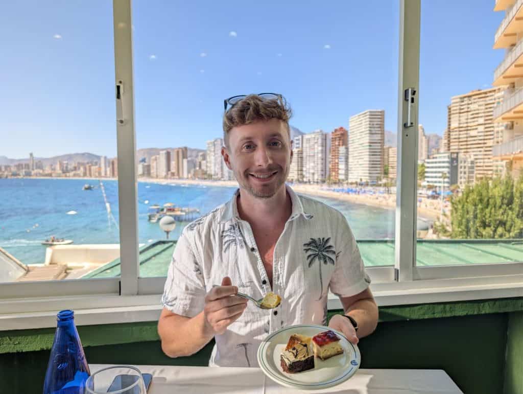 A smiling man enjoying a slice of cake at a seaside restaurant with a city skyline in the background.