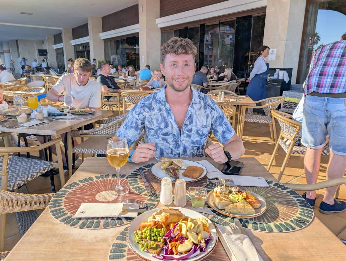 A young man enjoying a meal at an outdoor restaurant terrace with other diners in the background.
