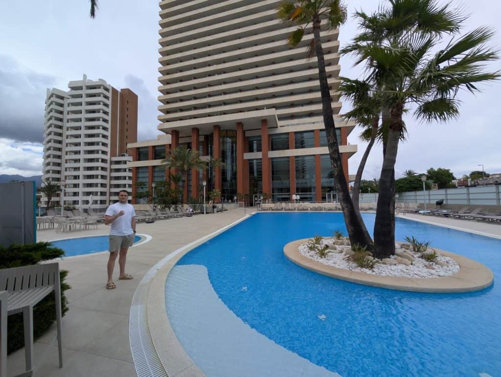 A lush hotel pool area with palm trees and modern high-rise buildings in the background.