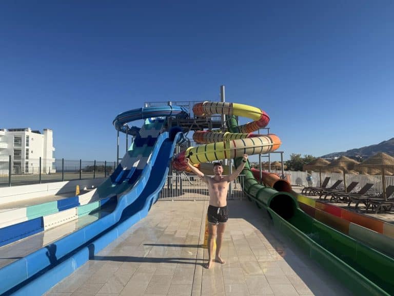 A man standing at the bottom of colorful water slides at a water park under a clear blue sky.