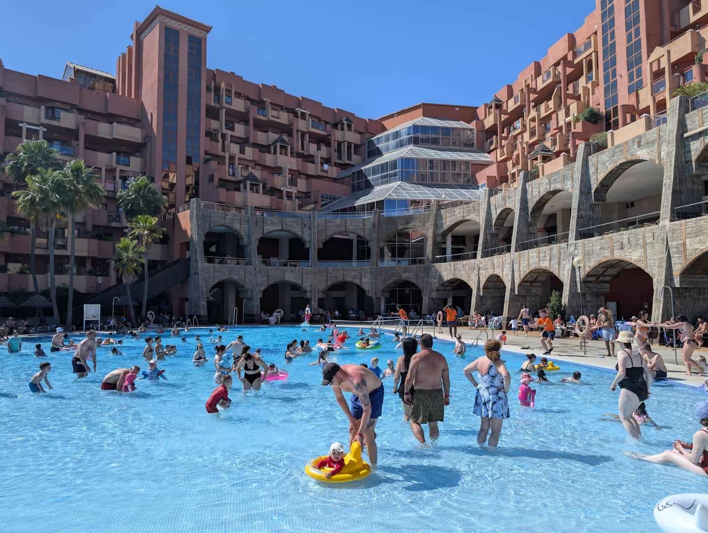 A lush hotel pool area filled with families and children enjoying the water in front of a large, mul.