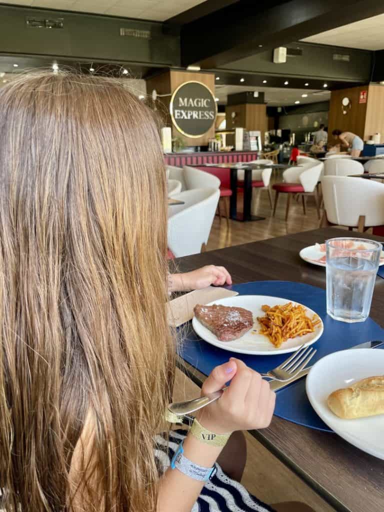 Niña comiendo un filete con pasta en una mesa del buffet del Magic Fantasy