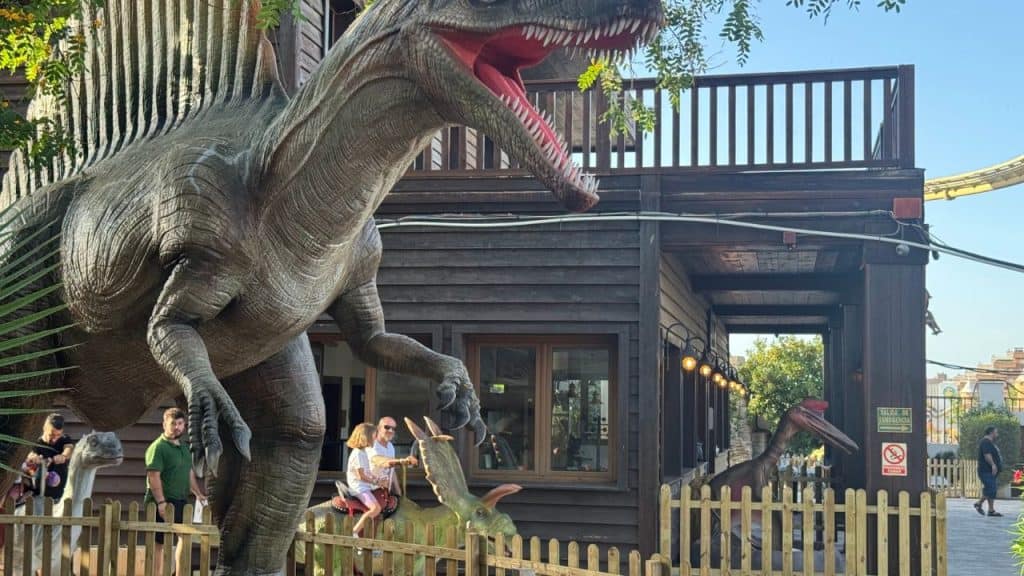 Dinosaur sculpture at a themed amusement park with visitors nearby.