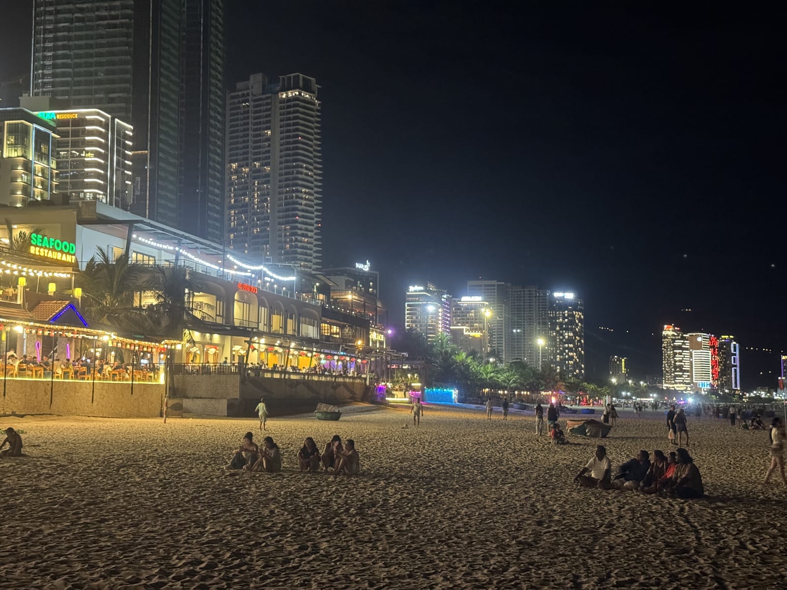 La playa de Da Nang de noche, con gente sentada en la arena y el paseo iluminado