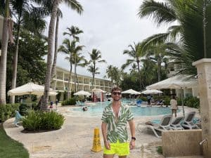 Man in tropical resort pool area with palm trees and lounge chairs.